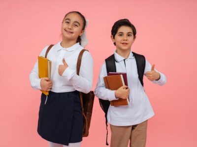 Two smiling schoolchildren give thumbs up with books and backpacks