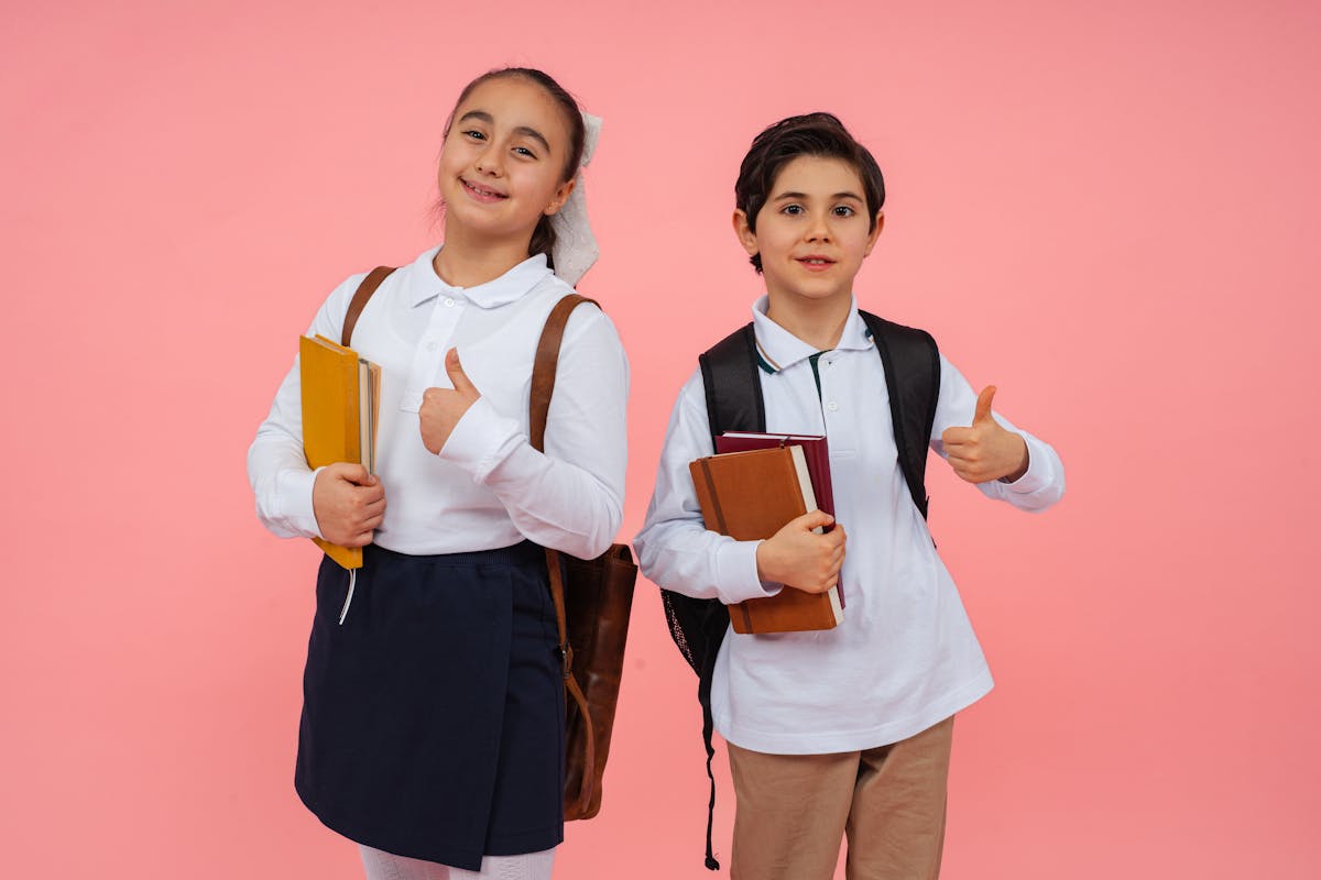 Two smiling schoolchildren give thumbs up with books and backpacks